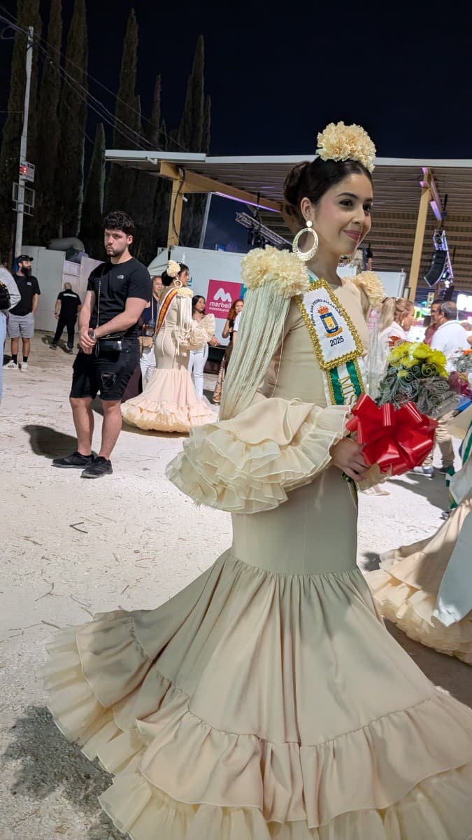 Feria de San Pedro 2025, 15 oct — joven con traje de flamenca y ramo, tradición y ambiente familiar en el recinto ferial de San Pedro Alcántara.