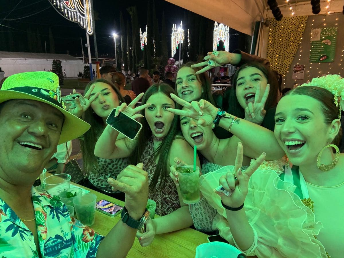 Feria de San Pedro 2025, 15 oct — grupo de amigos celebrando con copas y posando en una caseta iluminada en San Pedro Alcántara