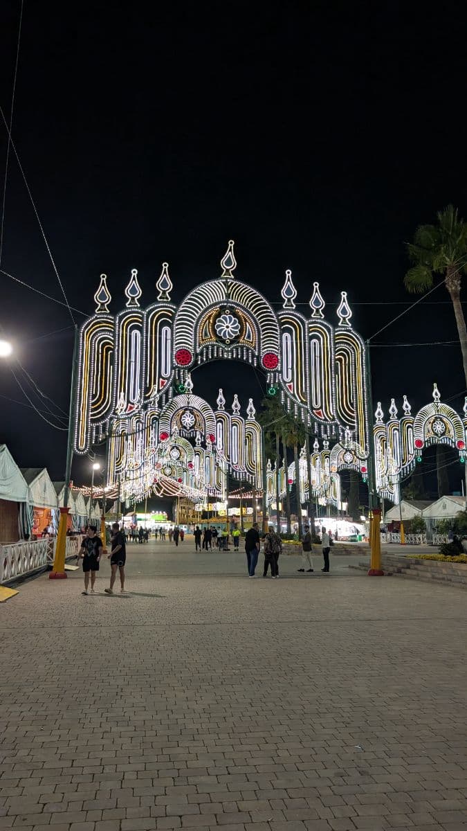 Feria de San Pedro 2025, 15 oct — portada principal iluminada del recinto ferial de La Caridad con visitantes paseando de noche.